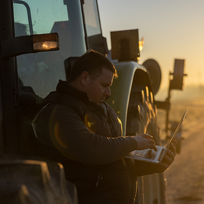 Farmer leaning on tractor using laptop.